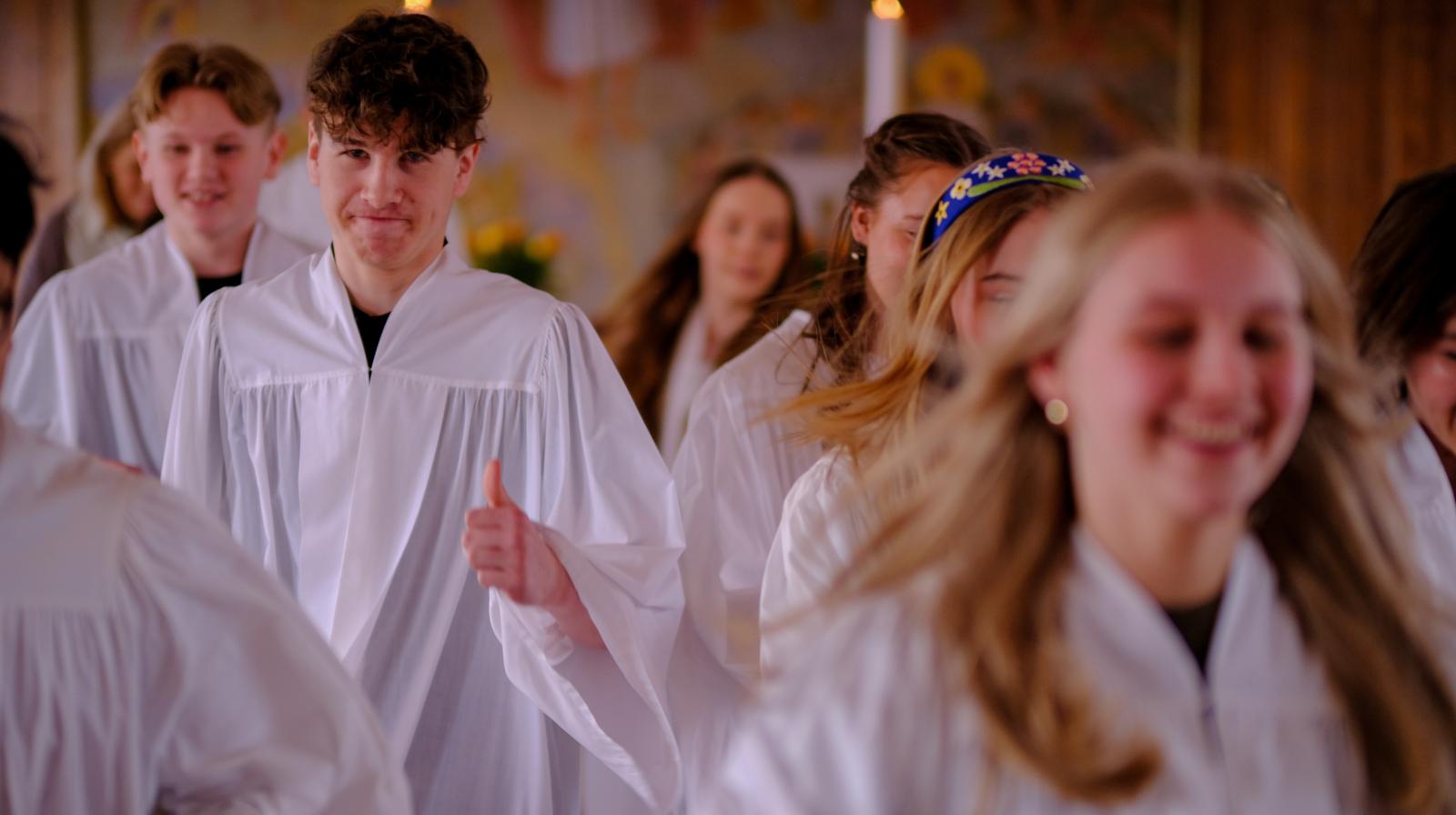 En gruppe konfirmanter i et kirkerom går mot kamera: Jenter i forgrunnen, og en gutt med tommel opp og smil til kamera i bakgrunnen.